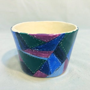 May include: A hand-painted ceramic planter with a patchwork design. The planter features a cream-colored interior and a vibrant exterior with geometric shapes in shades of blue, green, and purple, accented with white stitching.