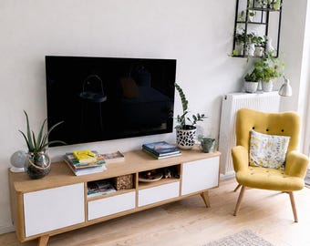 以下が含まれることがあります： 白いドアと引き出しが付いた、明るい木製のテレビ台があるモダンなリビングルーム。 黄色のアームチェアが台の隣にあり、黒い金属製の植物棚が壁に取り付けられています。 大きなテレビが台の上に設置されています。
