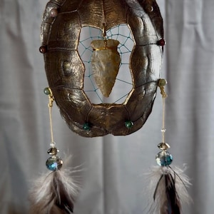 May include: A handcrafted dreamcatcher featuring a bronze-colored turtle shell frame. The center holds a web with a stone arrowhead. Decorative beads and feathers dangle below, creating a unique, rustic aesthetic.