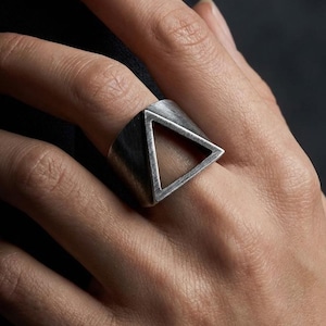 May include: A wide silver-toned ring featuring a prominent triangular cutout design. The metal surface has a brushed, matte finish with visible linear texture, creating a geometric aesthetic that wraps around the finger with a bold, open frame.