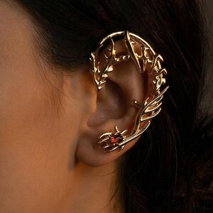 May include: A gold-toned ear cuff featuring an intricate, branch-like metal structure that wraps around the upper ear cartilage. The design includes a small, round, deep red gemstone set near the earlobe, creating a detailed, metallic accessory.
