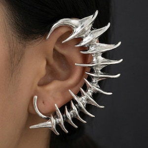May include: A silver-toned ear cuff featuring a curved, elongated spine-like structure with multiple sharp, protruding spikes that follow the natural contour of the outer ear, crafted with a metallic finish and textured detailing.