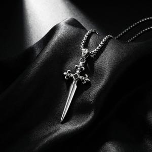 Puede incluir: Un colgante de metal plateado con forma de daga, con una guarda en forma de flor de lis y una pequeña piedra negra cuadrada en el centro. El colgante cuelga de una cadena de eslabones cuadrados a juego, con un acabado pulido y detalles intrincados.