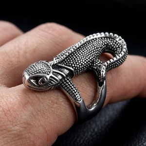 Puede incluir: Un anillo de metal plateado con forma de lagarto y superficie texturizada de escamas. La cabeza del lagarto mira hacia adelante, mientras que su cuerpo se curva alrededor del dedo para formar la banda, con escamas detalladas en relieve.