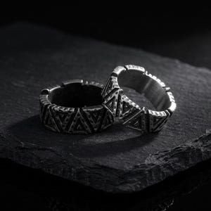 May include: Two silver-toned metal rings featuring a repeating geometric pattern of triangles around the outer band. The rings have a dark, oxidized finish with a textured, weathered appearance and a wide, flat profile design.