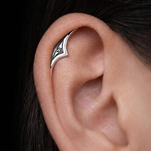 Puede incluir: Un pendiente tipo ear cuff de plata con un diseño curvo y puntiagudo que sigue el contorno del cartílago superior de la oreja. La superficie metálica tiene un acabado pulido con un símbolo de nudo triquetra grabado en un marco triangular.