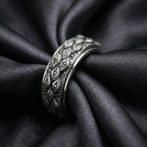May include: A silver-toned metal ring featuring a repeating pattern of embossed eyes across the entire outer band. The surface of the ring has a dark, oxidized finish within the recessed areas to highlight the detailed eye motifs and textures.