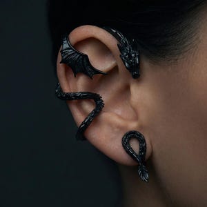May include: A matte black ear cuff shaped like a dragon, featuring a detailed head at the top, a textured wing extending across the upper ear cartilage, a winding scaly body wrapping around the ear, and a separate matching dragon tail stud.