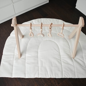 Op de afbeelding: Een witte gewatteerde speelmat met een houten speelgym-boogbrug met vier hangende houten speelgoedjes. De speelgoedjes omvatten een kralenring, een houten maan, een houten spiegel en een houten wolk.