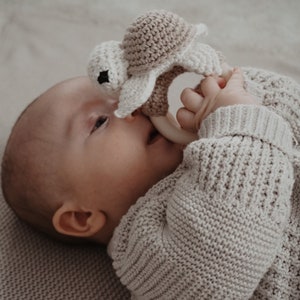 May include: A baby in a light grey sweater holds a crochet turtle teether. The teether is beige and white, with a wooden ring. The baby is lying on a light grey knitted blanket.