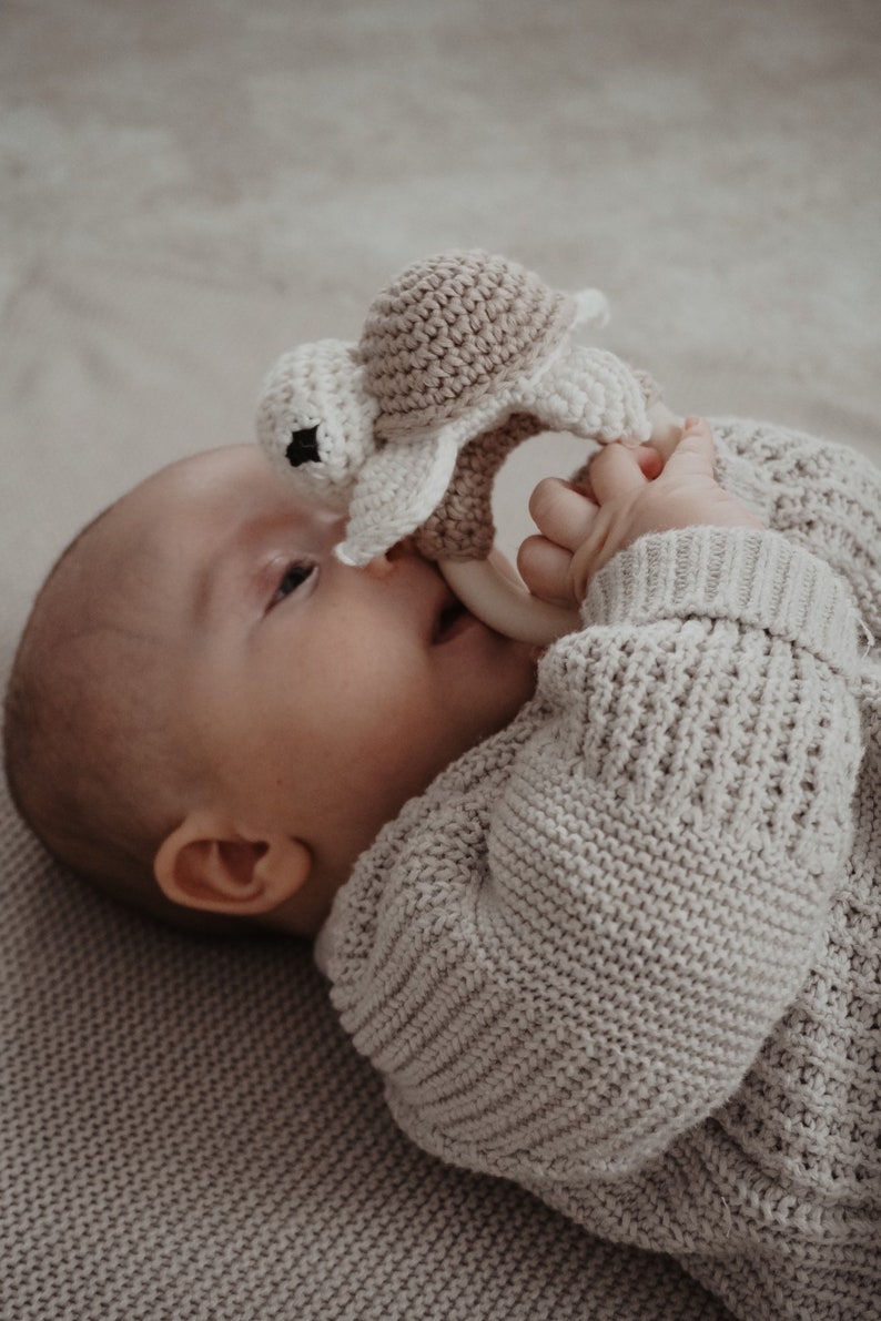 May include: A baby in a light grey sweater holds a crochet turtle teether. The teether is beige and white, with a wooden ring. The baby is lying on a light grey knitted blanket.