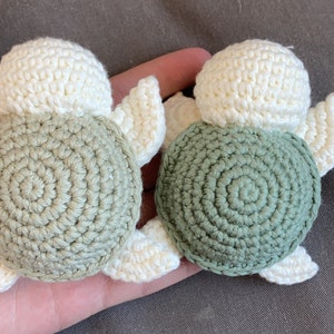 May include: Two handmade crochet turtle ornaments. Each turtle has a cream-coloured head and flippers, with a spiral-patterned shell. One shell is a muted green, the other is a darker green. The turtles are small, perfect for a nursery or as a gift.