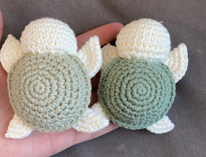 May include: Two handmade crochet turtle ornaments. Each turtle has a cream-coloured head and flippers, with a spiral-patterned shell. One shell is a muted green, the other is a darker green. The turtles are small, perfect for a nursery or as a gift.