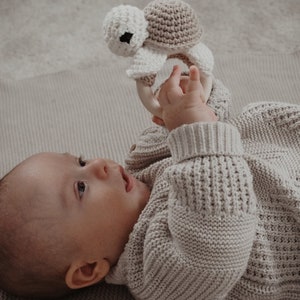 May include: A baby in a light grey knitted sweater looks upwards, holding a crochet turtle rattle. The rattle has a white head, a tan shell, and a wooden ring. The baby is lying on a matching grey blanket, creating a soft and gentle scene.