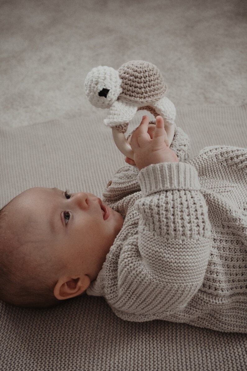 May include: A baby in a light grey knitted sweater looks upwards, holding a crochet turtle rattle. The rattle has a white head, a tan shell, and a wooden ring. The baby is lying on a matching grey blanket, creating a soft and gentle scene.