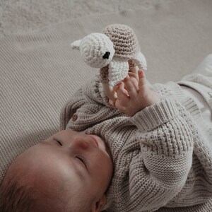 May include: A baby in a light grey knitted cardigan plays with a handmade crochet turtle toy. The turtle has a beige shell and a white head with black eyes. The baby lies on a light beige blanket.