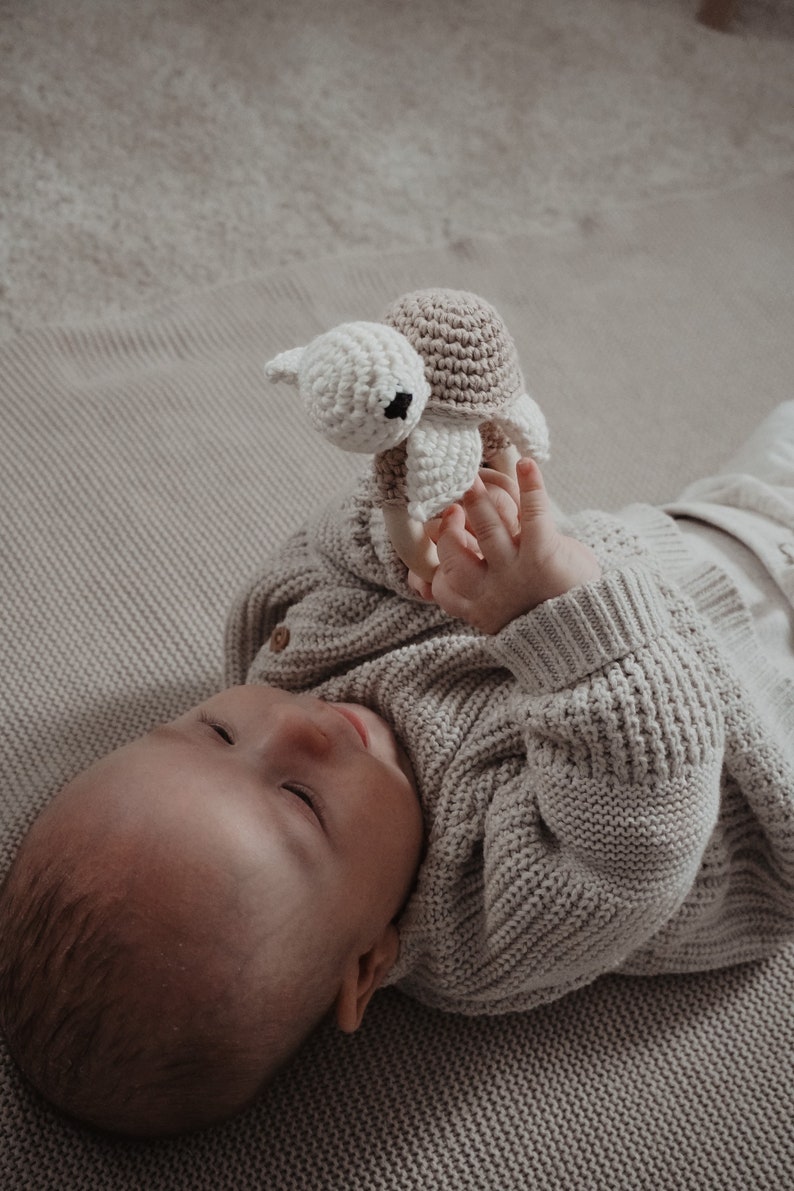 May include: A baby in a light grey knitted cardigan plays with a handmade crochet turtle toy. The turtle has a beige shell and a white head with black eyes. The baby lies on a light beige blanket.