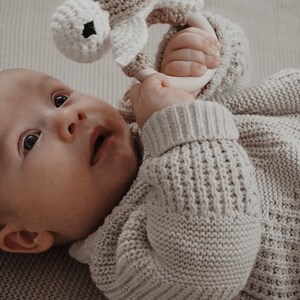 May include: A baby in a light grey knitted sweater holds a crocheted rattle. The rattle is designed as a turtle, made with beige and white yarn. The baby lies on a light grey textured surface.
