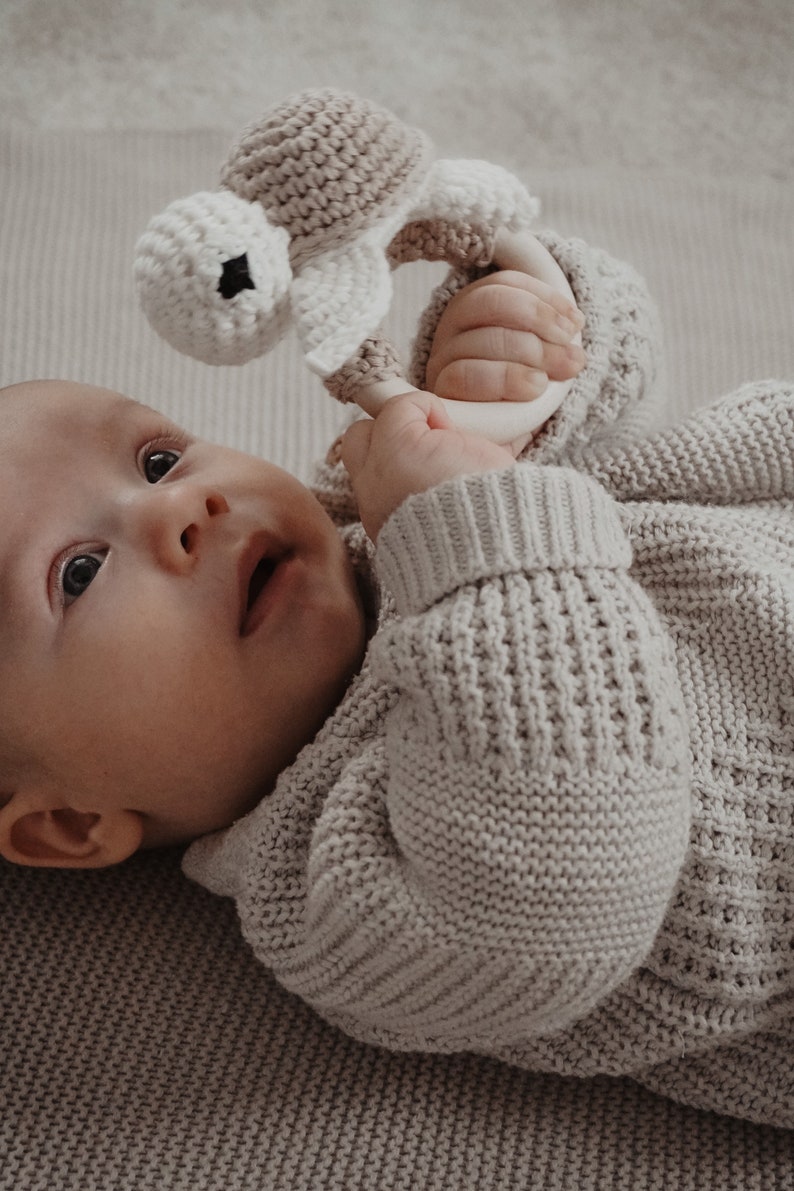 May include: A baby in a light grey knitted sweater holds a crocheted rattle. The rattle is designed as a turtle, made with beige and white yarn. The baby lies on a light grey textured surface.