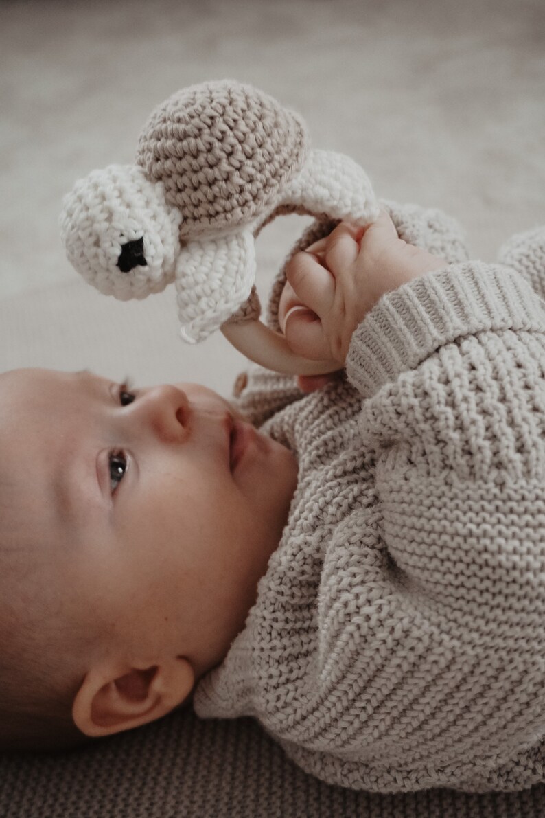 May include: A cream and tan crocheted turtle baby rattle. The turtle features a round head and shell, with a wooden ring. The rattle is designed for infants and toddlers.