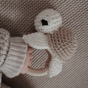 May include: A crocheted turtle baby rattle in cream and tan. The turtle has a cream-coloured head with black eyes and a tan shell. The rattle is held by a person wearing a cream-coloured sweater.