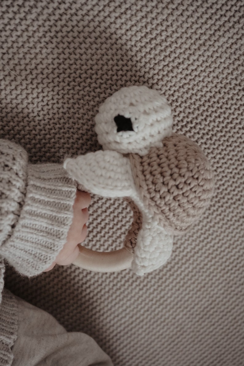 May include: A crocheted turtle baby rattle in cream and tan. The turtle has a cream-coloured head with black eyes and a tan shell. The rattle is held by a person wearing a cream-coloured sweater.