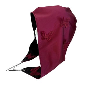 May include: A burgundy head covering with embroidered butterfly accents. The hood is lined with black fabric and features a drawstring closure. The design includes three embroidered butterflies on the exterior.