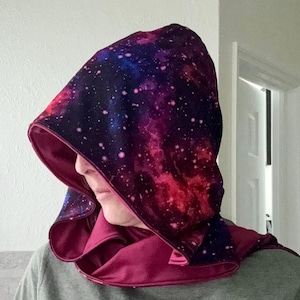 May include: A hooded cloak with a galaxy print in shades of purple, blue, and pink. The interior and trim are a solid burgundy color. The hood is draped over the shoulders.