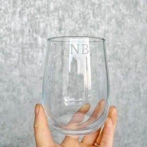 May include: A clear stemless wine glass with the initials "NB" etched on the front. The glass is held in a hand against a grey background. The glass has a rounded shape and is designed for wine.