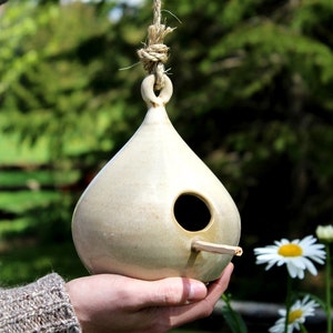 May include: A ceramic birdhouse shaped like a teardrop, painted in a light beige colour, with a small round hole for the birds to enter. The birdhouse is hanging from a natural twine rope and is being held by a hand.