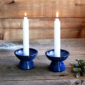 May include: Two white candles burning in blue ceramic candle holders on a wooden surface.