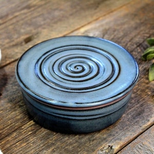 May include: A blue ceramic soap dish with a spiral design on the lid. The dish is sitting on a wooden surface.