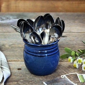 May include: A blue ceramic jar filled with silver spoons. The jar is sitting on a wooden surface with a white and gray striped cloth napkin and a sprig of white flowers.