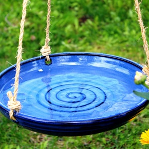 May include: A blue ceramic bird bath with a spiral design hangs from a rope in a grassy garden.