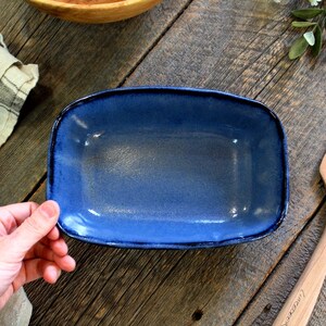 May include: A blue rectangular ceramic baking dish with a dark blue rim. The dish is empty and sitting on a wooden surface.