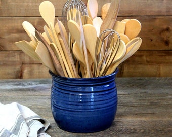 May include: A blue ceramic utensil holder filled with wooden spoons and whisks. The holder has a textured, glazed finish.