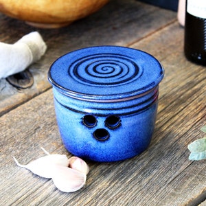 Peut inclure: Un pot à ail en céramique bleu avec un couvercle orné d'un motif en spirale. Le pot possède trois trous d'aération et est posé sur une surface en bois avec des gousses d'ail et des tomates.