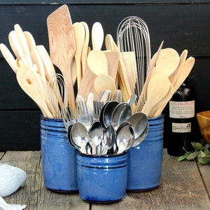 Peut inclure: Trois porte-ustensiles en céramique bleue contenant une sélection de cuillères en bois, de spatules et d'ustensiles en métal. Les porte-ustensiles sont sur une surface en bois.