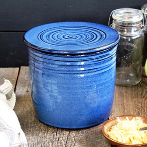 Peut inclure: Un pot en céramique bleu avec un couvercle, utilisé pour fermenter des aliments comme la choucroute. Le pot est sur une surface en bois avec un bol de chou râpé et deux bocaux en verre.