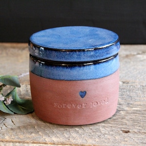 May include: A blue and brown ceramic jar with a lid. The jar has the words "Forever loved" and a small heart engraved on the side.