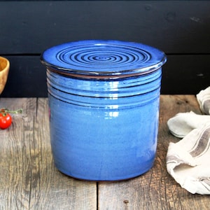 May include: A blue ceramic jar with a lid. The jar has a swirl design on the lid and a glazed finish. The jar is sitting on a wooden surface.