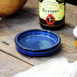May include: A small blue ceramic dish with a black rim. The dish has a swirl pattern on the inside. The dish is sitting on a wooden surface with a clove of garlic and some tomatoes.