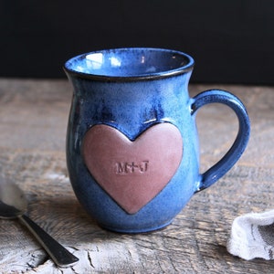 May include: A blue ceramic mug with a heart-shaped design. The heart is a light pink colour and has the initials "M+J" engraved on it.