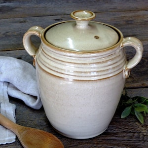 May include: A large, off-white ceramic crock with a lid and two handles. The crock has a glazed finish and is decorated with horizontal lines.