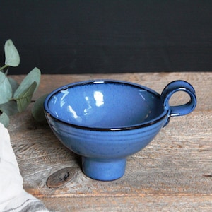 May include: A small blue ceramic bowl with a handle and a pedestal base. The bowl has a black rim and a glossy finish.