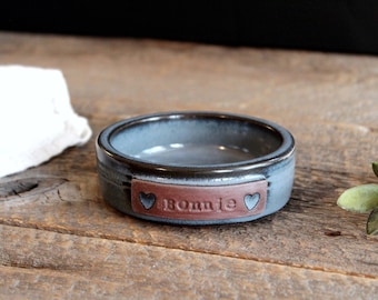 May include: A small, blue ceramic pet bowl with a brown leather tag that says "Bonnie" with two hearts on either side.