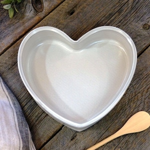 May include: A white heart-shaped ceramic bowl on a wooden surface. The bowl is empty and has a smooth, glossy finish.