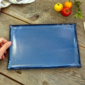 Peut inclure: Une assiette rectangulaire en céramique bleue avec un bord légèrement surélevé. L'assiette est posée sur une surface en bois avec deux paires de baguettes et trois tomates rouges.