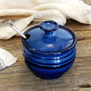 May include: A blue ceramic sugar bowl with a lid and a spoon resting on the rim. The bowl has a dark blue glaze and a slightly textured surface.