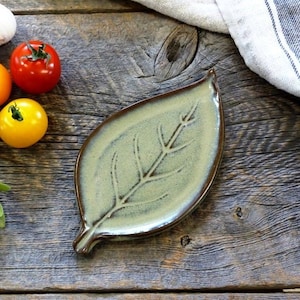 Peut inclure: Un plat en céramique en forme de feuille avec un glaçage vert clair et une bordure marron. Le plat est posé sur une surface en bois rustique, accompagné de tomates, d'ail et d'une brindille de verdure. Un tissu rayé est en arrière-plan.